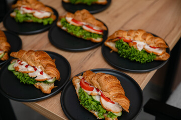Delicious croissant sandwiches with lettuce, tomato, cucumber, chicken, and sauce on black plates, served on a wooden table. Perfect for breakfast, brunch, or catering presentations.