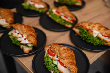 Delicious croissant sandwiches with lettuce, tomato, cucumber, chicken, and sauce on black plates, served on a wooden table. Perfect for breakfast, brunch, or catering presentations.