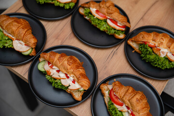 Delicious croissant sandwiches with lettuce, tomato, cucumber, chicken, and sauce on black plates, served on a wooden table. Perfect for breakfast, brunch, or catering presentations.