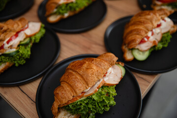 Delicious croissant sandwiches with lettuce, tomato, cucumber, chicken, and sauce on black plates, served on a wooden table. Perfect for breakfast, brunch, or catering presentations.