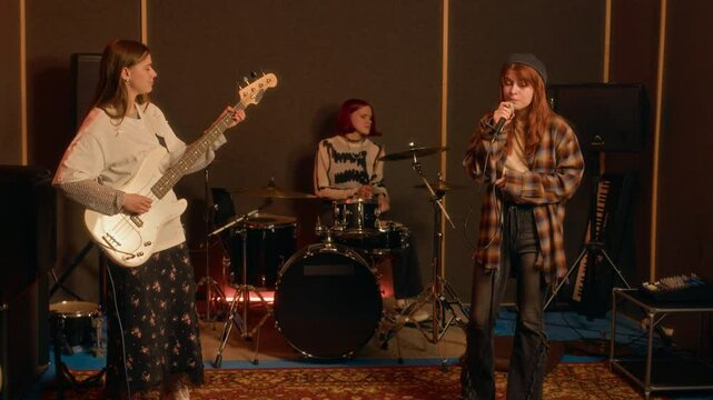 Three Gen Z girls playing in punk rock band, singing, playing drums and bass guitar, rehearsing in dark soundproof music studio