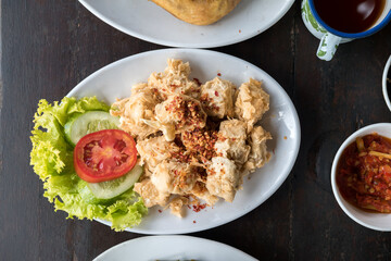 Crispy fried tofu (tahu goreng) sprinkled with chili flakes, served with lettuce, cucumber, and tomato on a white plate. A popular Indonesian side dish.