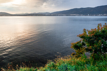A calm lake with a tree on the shore. The water is a deep blue color. The sky is cloudy and the sun is setting