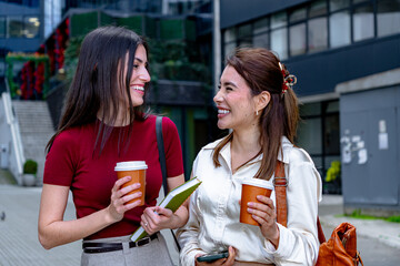 Two female professionals enjoying a conversation and holding takeaway coffee cups while walking outdoors near their office building