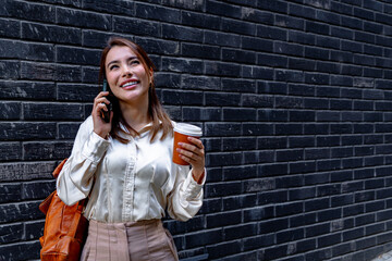 Young manager smiling while talking on smartphone and holding takeaway coffee in front of dark...