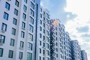 The buildings have a modern design with white and gray facades and many windows. Air conditioning is installed on each window. The sky is blue with small clouds. A glare of light from the sun is
