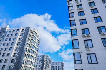 Fototapeta premium The photo shows a view from below of modern high-rise buildings against the background of a bright blue sky with white clouds. The buildings have white and gray facades, many windows, as well as air