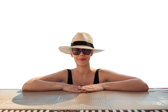 Girl in hat is in pool, leaning on side. Young woman with glasses and hat on vacation is relaxing on edge of pool on sunny day. Portrait of woman in pool looking at camera. Sitting on edge of pool