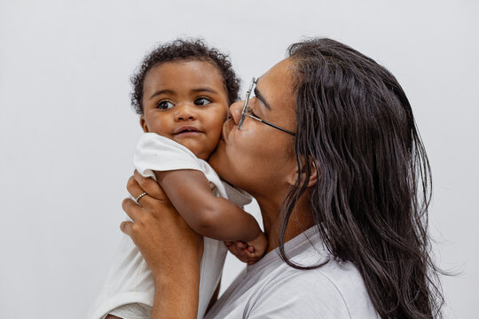 Beautiful Brazilian mother hugs and kisses her baby in the studio on a white background. A young mother in a white bodysuit hugs and plays with her baby in the studio. Black baby, African baby. Brazil