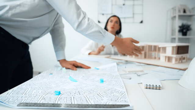 Skilled caucasian interior designer working with house model while discussing with asian architect engineer about house plan on meeting table with blueprint and house model scatter around. Immaculate.