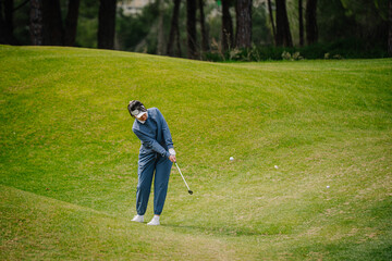 A female golfer in blue sportswear swings her club on a grassy hill during a golf game, surrounded by lush green fairway and trees in the background..