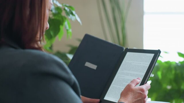 Mature woman using finger to scroll or turn page on digital e-reader while enjoying quiet reading moment indoors surrounded by green houseplants and soft natural light in peaceful cozy home setting