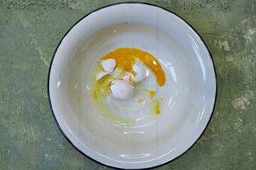 in a white enameled bowl on a green background is a broken egg with shell, top view