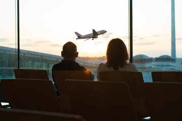 People wait in an airport lounge, silhouetted against a large window at sunset. The warm glow of...