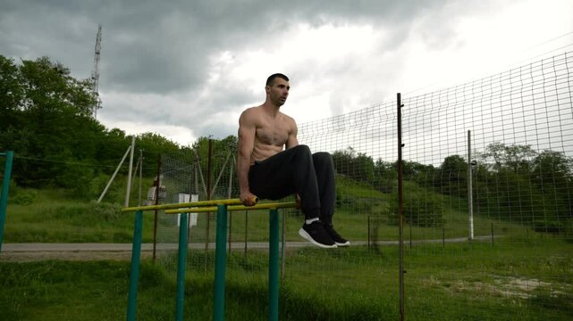 Strong guy training abdominal muscles on parallel bars at sports ground. Young shirtless sportsman exercising at nature. Sporty athlete working out at nature. Concept of active sportive lifestyle