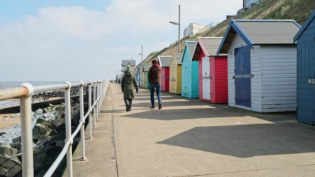 Unidentifiable couple walking along the promenade and colourful beach huts in the seaside town of Sheringham on the North Norfolk coast