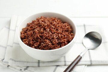 Tasty brown rice in bowl and spoon on white table, closeup