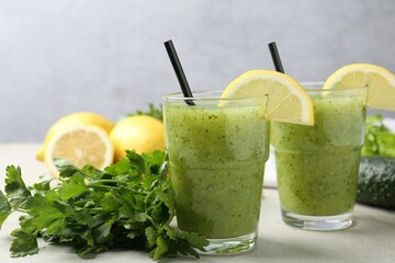 Healthy parsley smoothie in glasses, leaves and lemon on light textured table against grey background, closeup