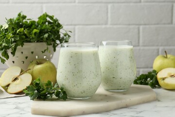Healthy parsley drink in glasses, leaves and apples on white marble table, closeup