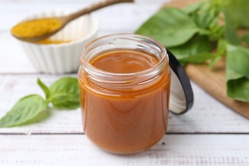 Tasty curry sauce in glass jar, powder and basil on white wooden table, closeup