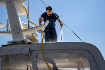 Luxury yacht being cleaned by a professional worker at the dock. Boat maintenance service in action at a marina with a bright summer sky and moored vessels.