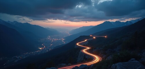 Winding road illuminated by car light trails at dusk. Serpentine route leads through mountain landscape toward cityscape lights. Travel adventure exploration concept. Dark sky above.