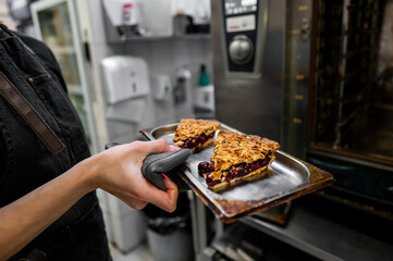 A chef wearing an apron carefully removes a tray of freshly baked berry pie slices from the oven,...