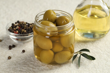 Pickled green olives in jar, oil and peppercorns on white textured table, closeup