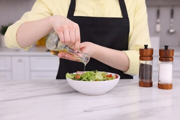 Woman pouring oil onto tasty salad at white marble table in kitchen, closeup