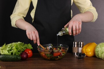 Woman pouring oil onto tasty salad at wooden table against dark background, closeup