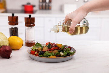 Woman pouring oil onto tasty salad at white marble table in kitchen, closeup