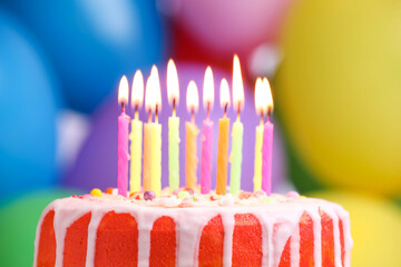 Delicious birthday cake with burning candles against colorful balloons, closeup