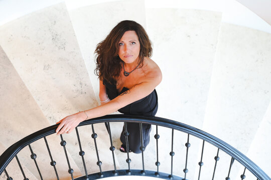 Beautiful woman walking up a spiral marble staircase looking up in an elegant setting. Her outfit features bare shoulders and her hand is resting on the railing. 