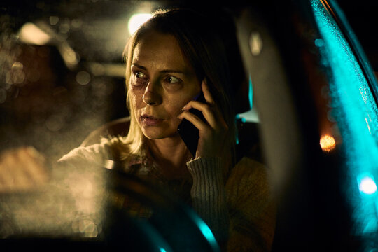 A young Caucasian woman, bearing the distressing signs of domestic abuse with prominent bruises and blackened eyes, speaks on her phone as she navigates her car through the nighttime streets.