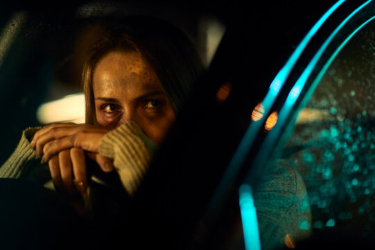 A young Caucasian woman, marked by the harrowing evidence of domestic abuse with distinct bruises and darkened eyes, sits in her car, leaning heavily on the steering wheel.