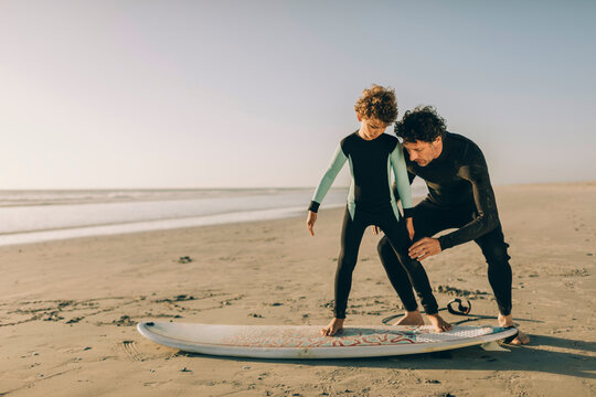 Young father teaching his son how to surf and become a surfer on a sandy beach while wearing surf suits