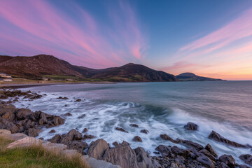 Fototapeta premium Rocky irish coastline at sunset with waves crashing onto shore, colorful pink clouds in blue sky and rolling hills in background
