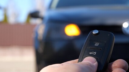 Man is pressing a button on a car's remote control, with the vehicle in the background out of focus. The car's right front blinker is blinking