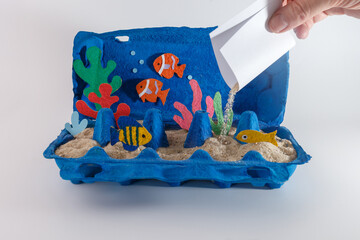 Creative underwater diorama project using a blue egg carton, sand, and colorful paper fish. A hand pours sand to complete the ocean floor. summer activities