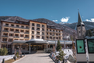 A multi story hotel with balconies and a glass canopy stands near a church tower with a clock face,...