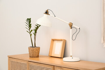Wooden chest of drawers with lamp, houseplant and frame near light wall in room