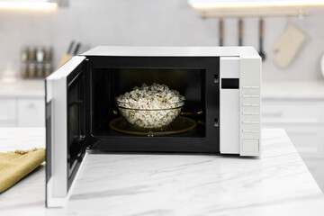 Open microwave oven with bowl of popcorn on white marble table in kitchen
