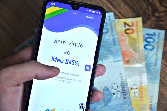 National Institute of Social Security. INSS. The app called Meu INSS on the screen of a cell phone that a man is holding and Brazilian currency notes in the background. Rio de Janeiro, Brazil-04.11.25