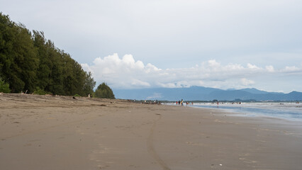 A scenic view of a sandy beach with people enjoying the waves, backed by mountains and a cloudy sky.