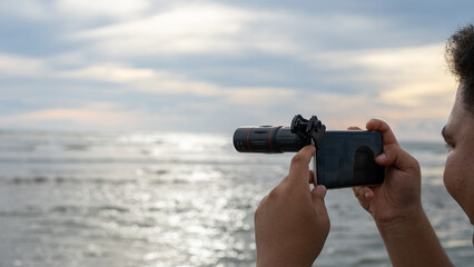 Obraz premium A person using a smartphone with a telephoto lens attachment to capture the ocean view during sunset.
