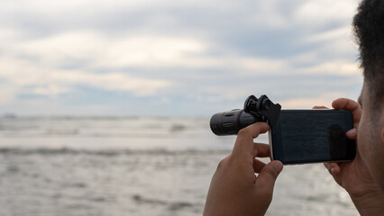 Obraz premium A person captures the seascape using a smartphone with a telephoto lens attachment. The blurred ocean horizon and cloudy sky create a serene atmosphere, highlighting mobile photography's creativity.