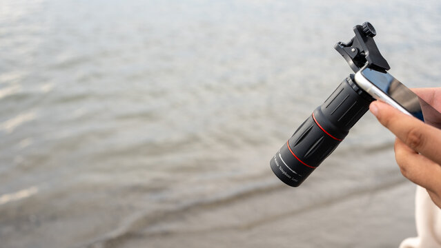 A close-up of a smartphone with a telephoto lens attachment, held over calm seawater. The compact clip-on lens enhances mobile photography, showcasing innovation in portable optics.