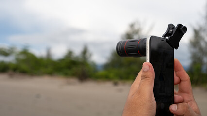 A person using a smartphone with a clip-on telephoto lens to capture a distant subject on a beach....