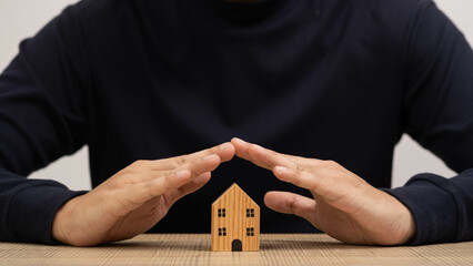 A person is safeguarding a miniature wooden house with hands, symbolizing real estate investment...