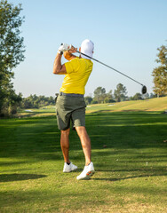 A golfer wearing a yellow shirt taking a swing at a golf ball down a fairway on a sunny day